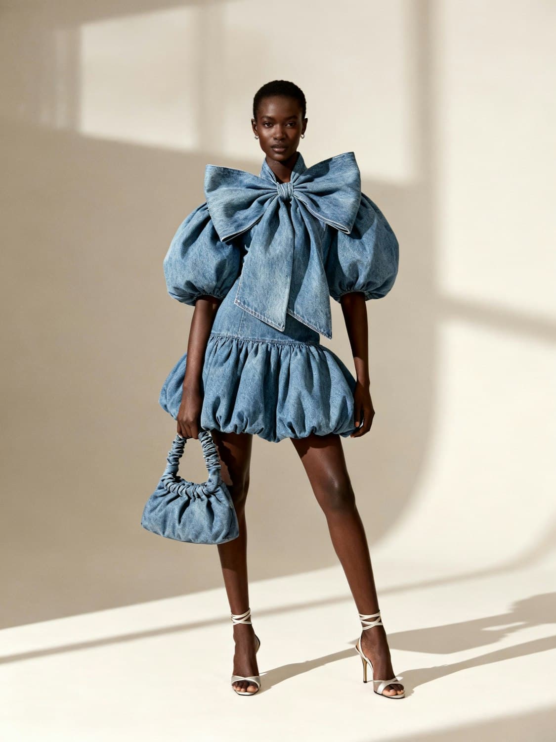 Model in an oversized denim bow dress with puffed sleeves holding a matching gathered denim bag.