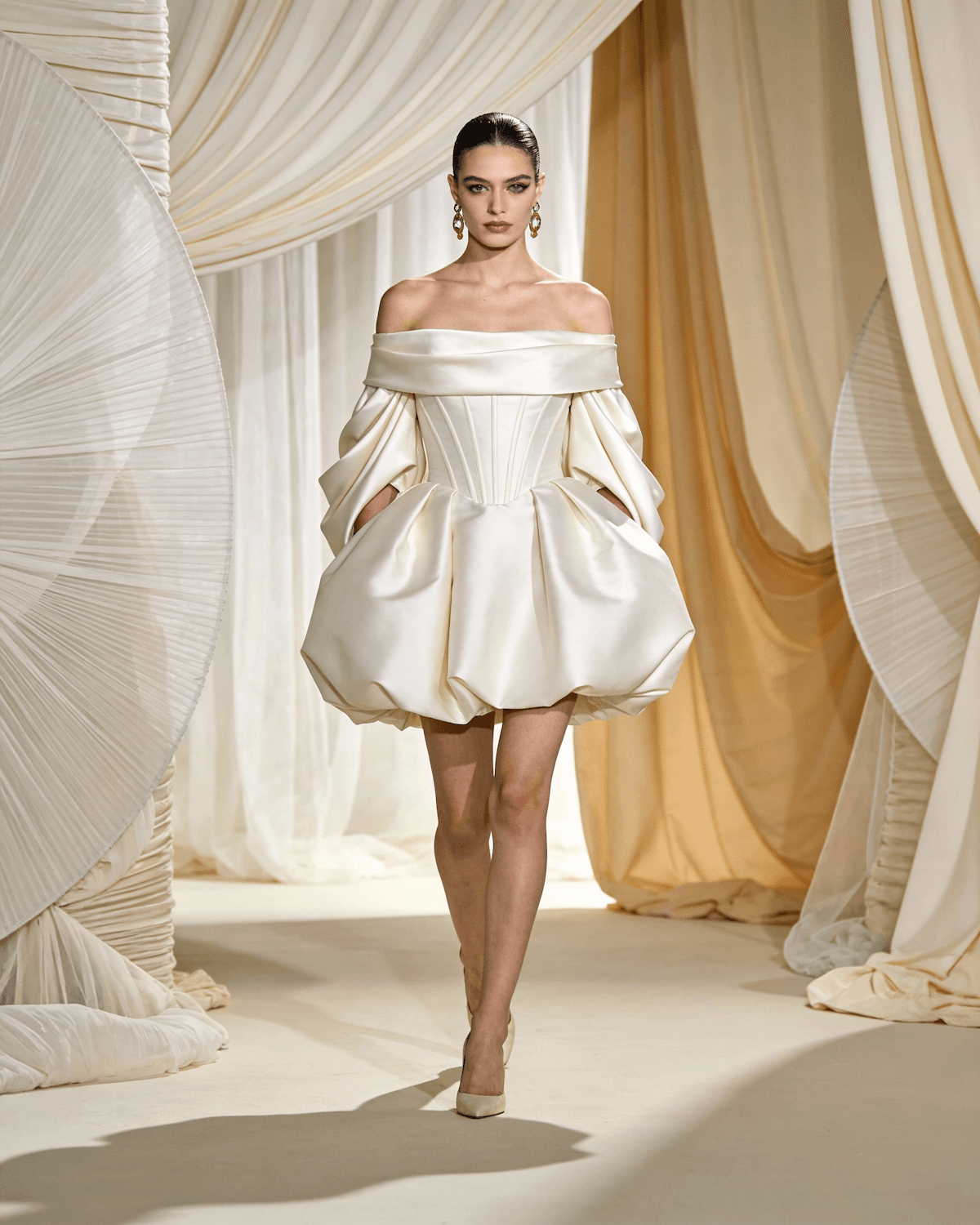 Model in an off shoulder ivory mini dress walking a runway framed by cream drapery and giant pleated fans.