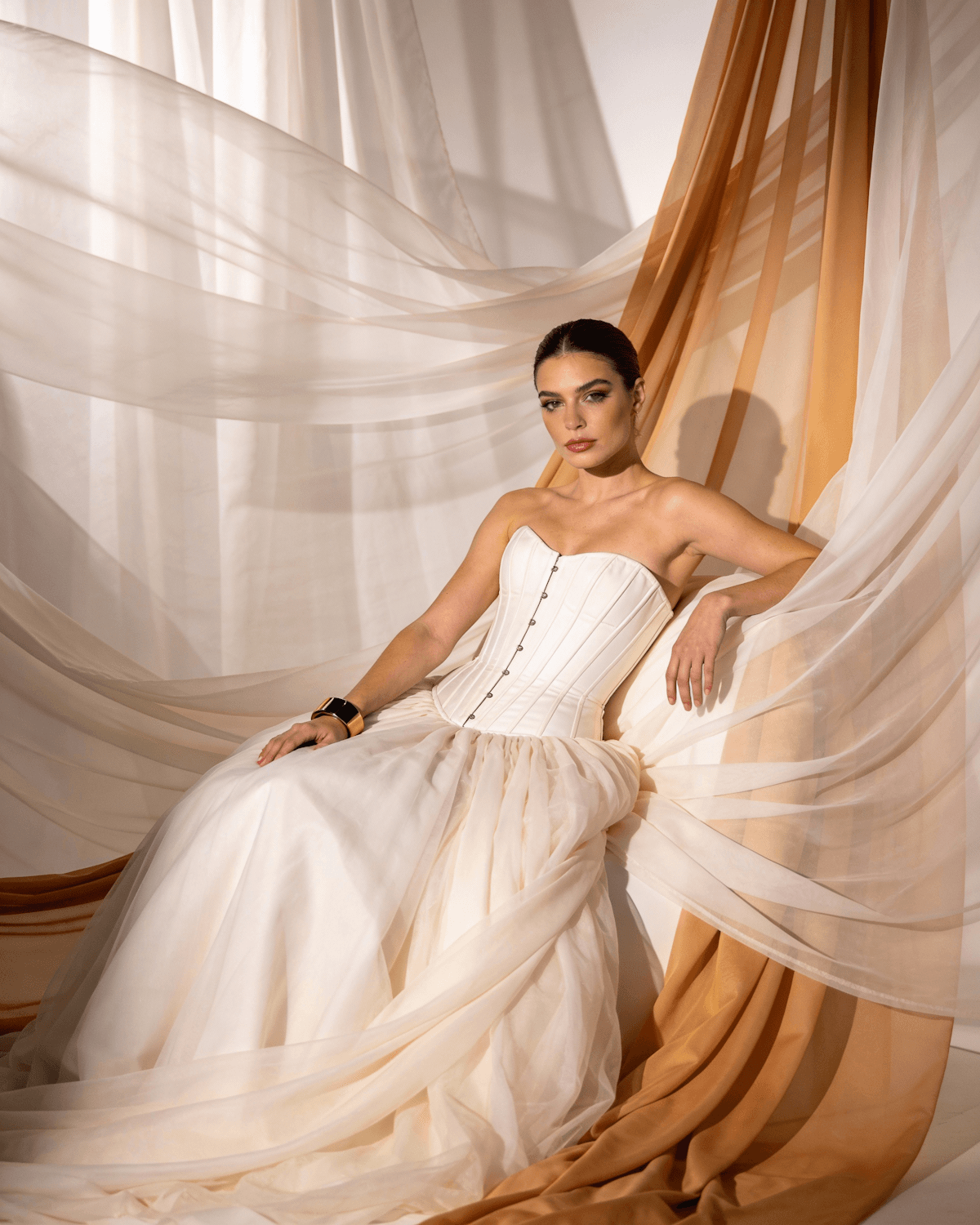 Woman in a strapless ivory corset gown seated among sweeping sheets of cream and caramel chiffon.